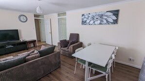 A living room with a sofa, a TV, a dining table with chairs, a chandelier, and a door. - A&A Apartment Basildon with free parking  (Basildon)