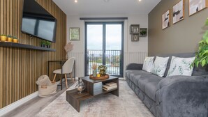 Una sala moderna con un sofá gris, un televisor de pantalla plana montado en una pared de madera, un ventanal con vistas y una mesita auxiliar con objetos decorativos.