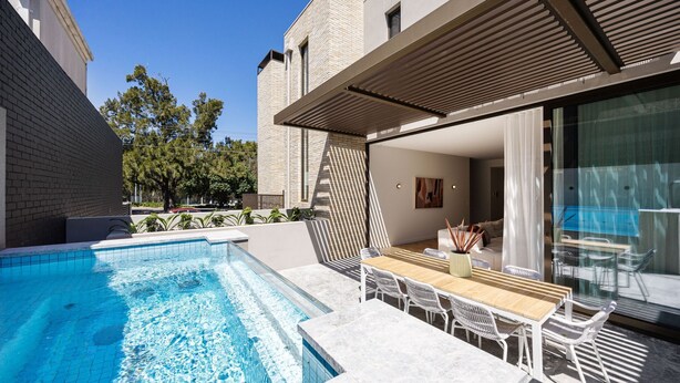 Un área de piscina moderna al aire libre con una mesa y sillas de madera, una puerta de cristal que da a una sala de estar y un edificio de ladrillo al fondo.