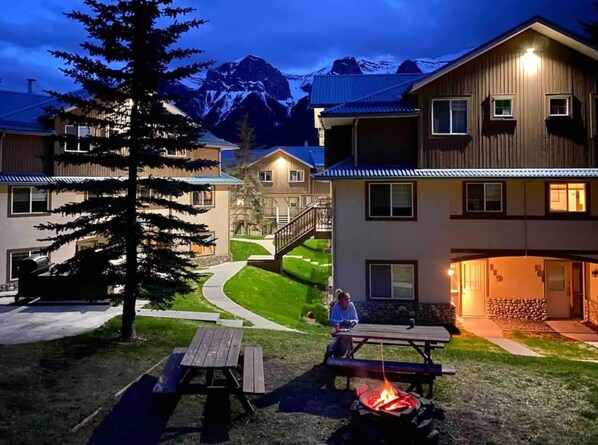A lodge with a fire pit and picnic table in the yard, surrounded by buildings and a mountain backdrop.