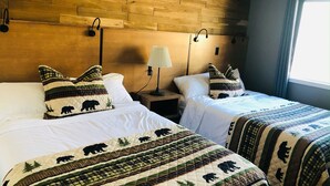 Ett hotellrum med två sängar, var och en med ett vadderat sängöverkast med björnmotiv, ett träfärgat sänggavel, sänglampor och ett fönster med gardiner.