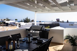 An outdoor patio with a grill, dining table, chairs, and a view of the sea.