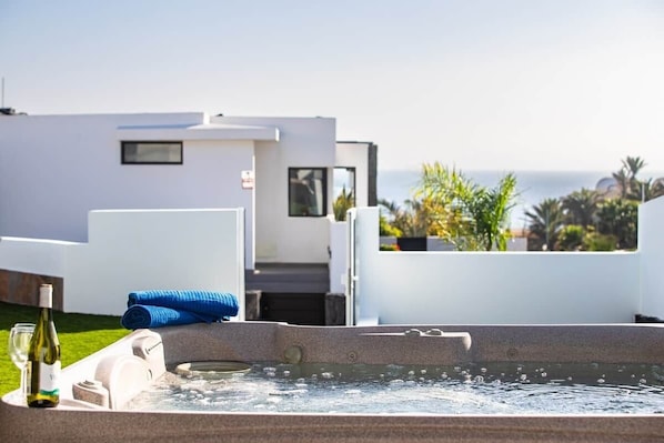 A modern outdoor hot tub with a clear view of the ocean and a white building in the background.