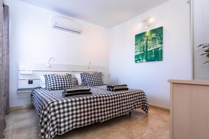 Una cama bien hecha con colcha a cuadros, una mesita de noche con lámpara y un aire acondicionado instalado en la pared.