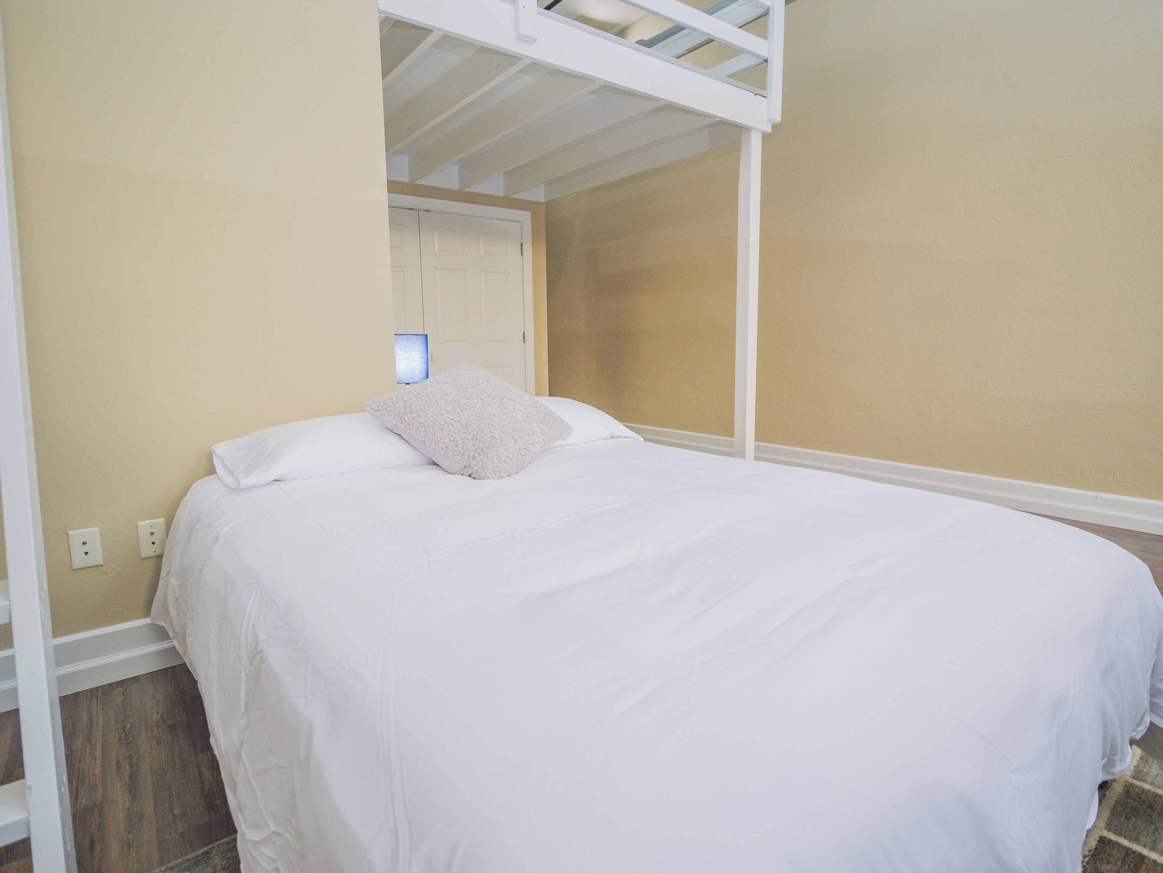 A bedroom with a bunk bed, white bedding, a bedside lamp, and a door in the background.