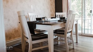 A dining table with four white chairs, a black table runner, and a window with a view of trees.