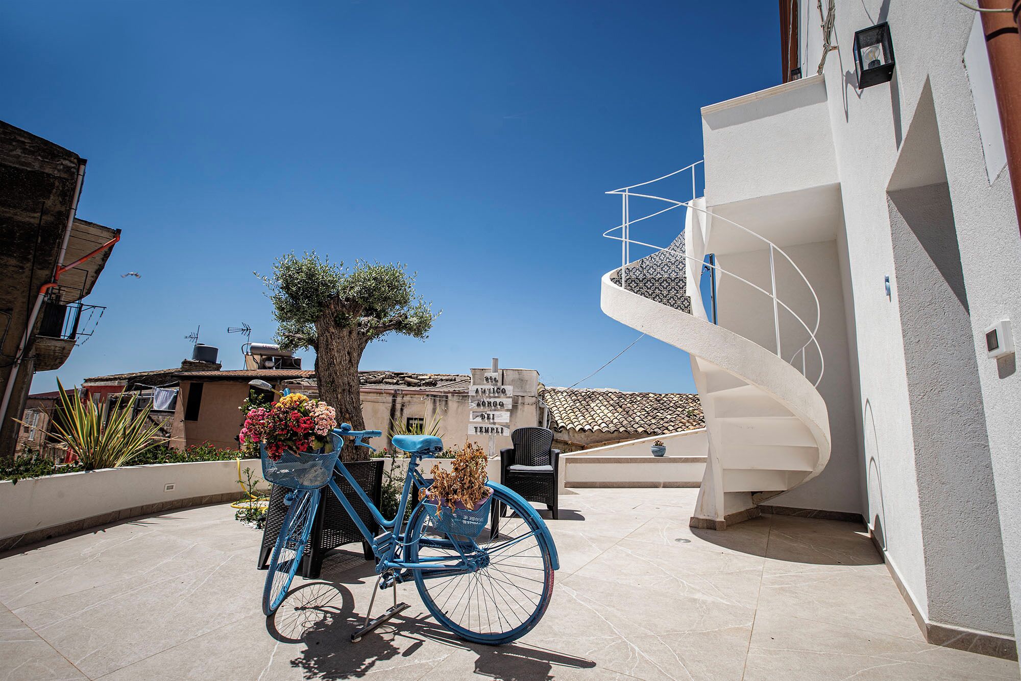 A blue bicycle with baskets, a tree, and a white building with a spiral staircase.