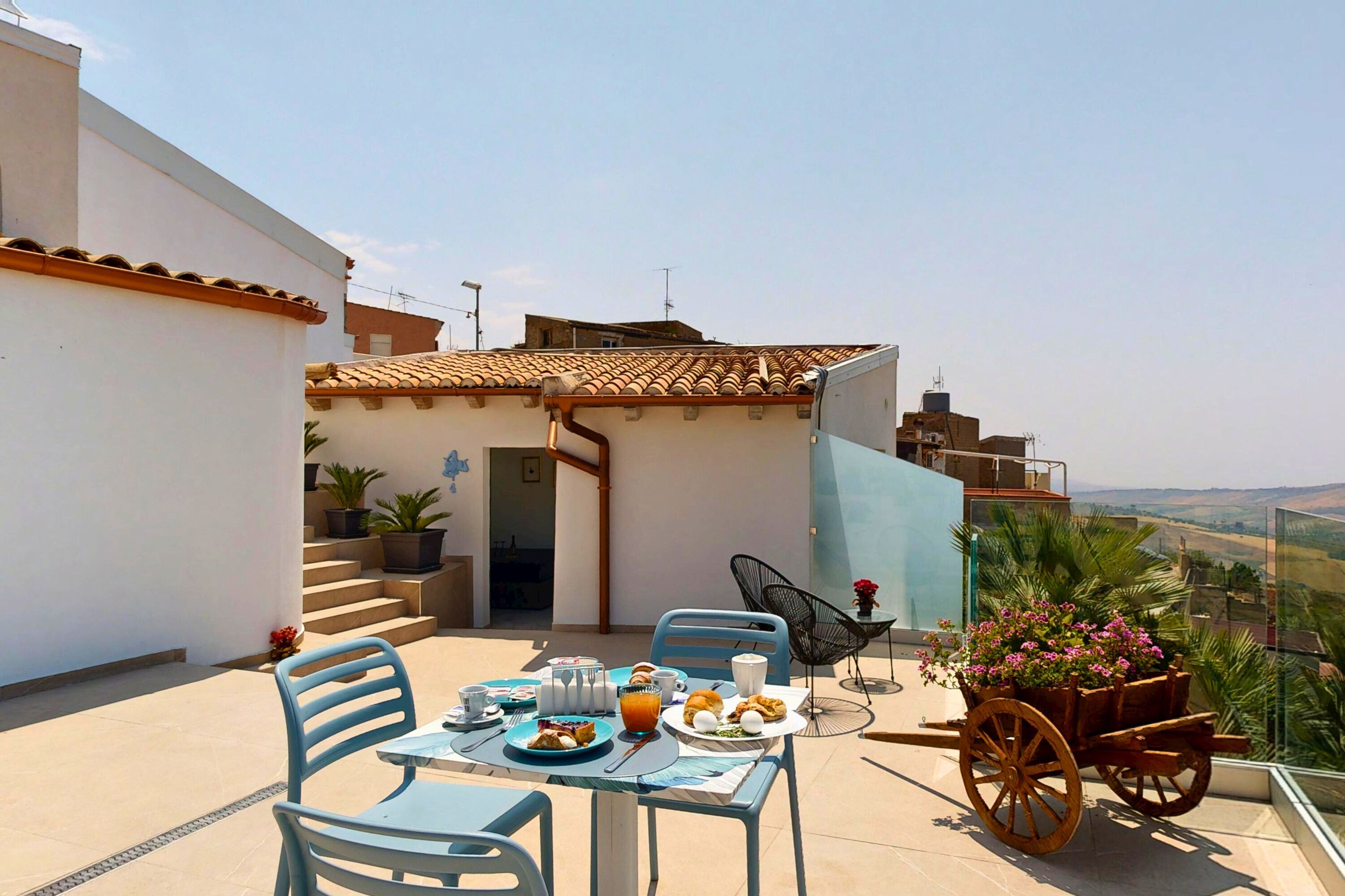 Una terraza en la azotea con una mesa lista para el desayuno, sillas, un carrito de madera con flores y vistas al paisaje.