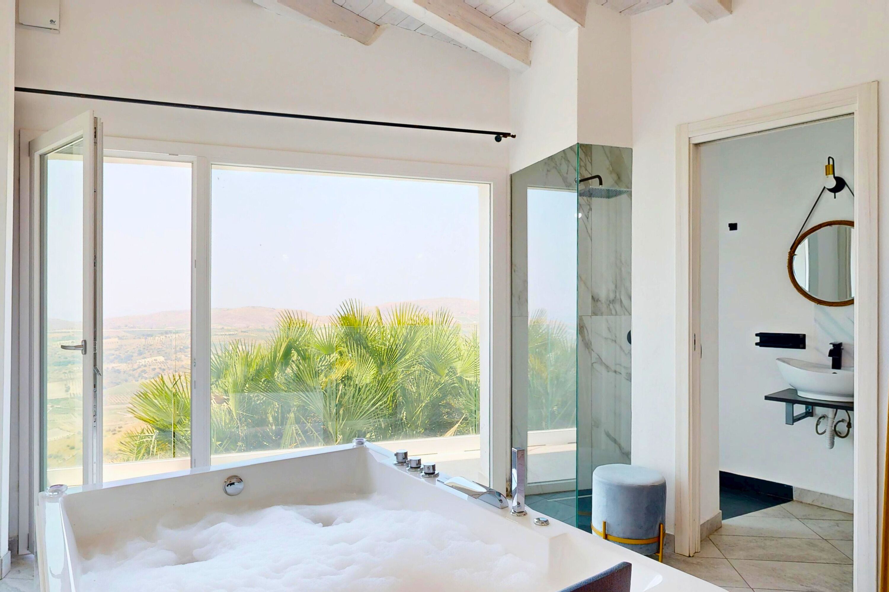 Ein modernes Badezimmer mit einer großen Badewanne, einer Duschkabine aus Glas und Blick auf Palmen.