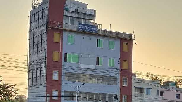 Un immeuble à plusieurs étages orné d’enseignes commerciales, une rue animée avec des piétons et un pousse-pousse, et un ciel dégagé.