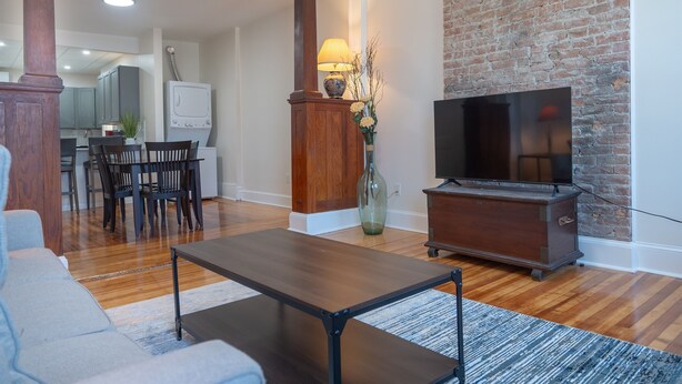 A living room with a wooden coffee table, a TV stand, and a brick wall. - Ironworks Urban Retreat|Pet Friendly|RPI|Hospitals (Troy)
