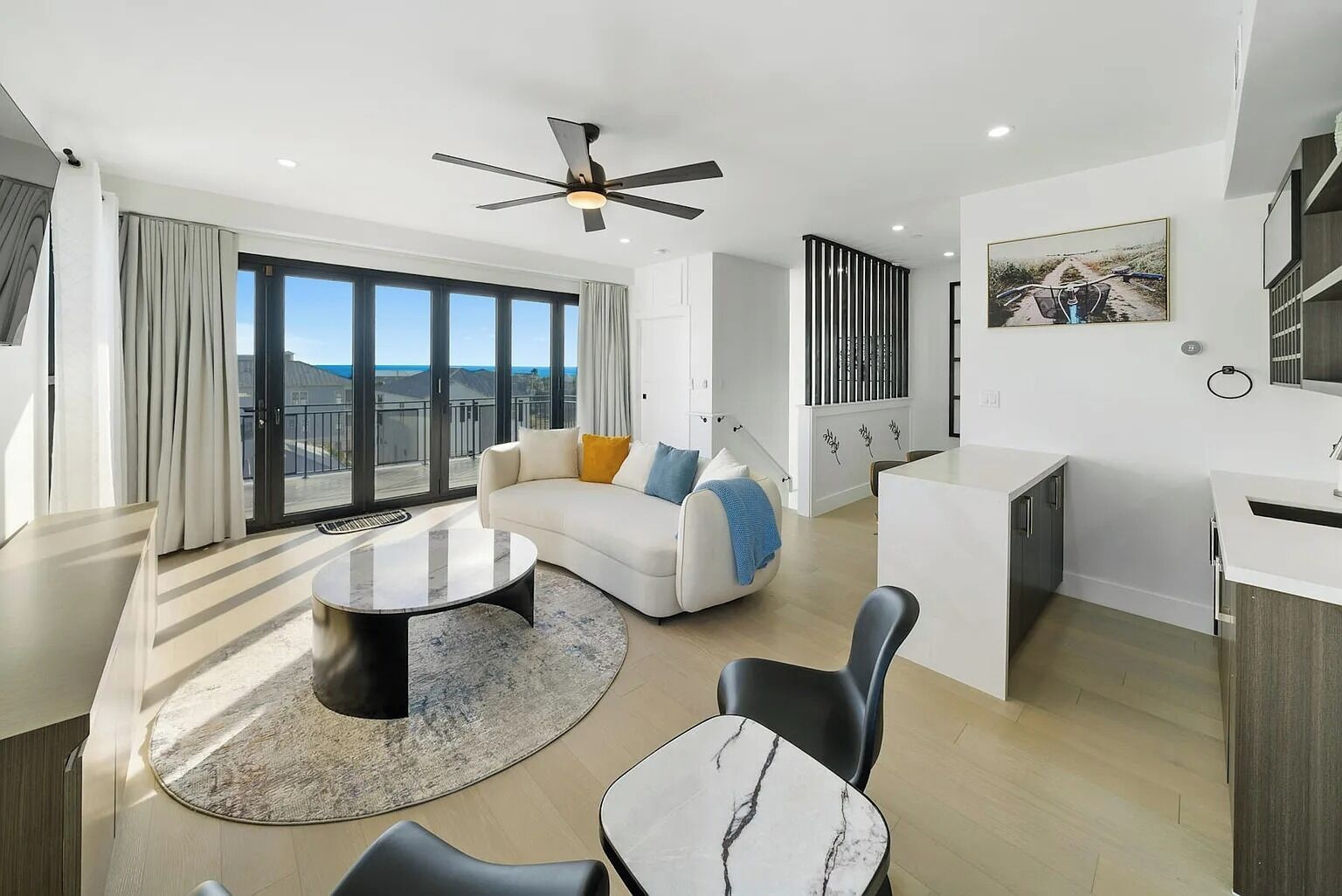 A modern living room with a sofa, coffee table, and chairs, a kitchen area, and large windows offering a view.