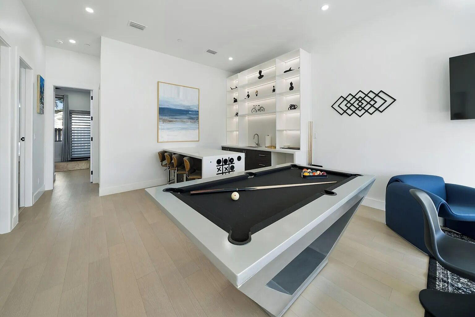 A modern pool table in a well-lit room with a minimalist design, a seating area with a blue chair, and a wall-mounted shelf with various items.