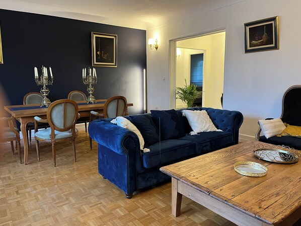 A living room with a dark blue sofa, wooden dining table, and chairs. There are two framed pictures on the walls and a potted plant in the corner.