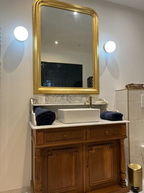 A bathroom with a marble countertop, a gold-framed mirror, wooden cabinets, and a gold trash can.