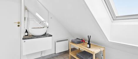 A modern bathroom with a skylight, a white sink, and a wooden shelf with towels and toiletries.