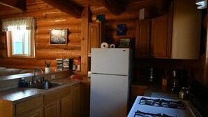 A log cabin kitchen with wooden cabinets, a white refrigerator, a gas stove, and a window with a view. - The Loon's Nest Cabin (Trapper Creek)