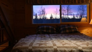 A bedroom with a large window, a bed with a patterned quilt, and a lamp on the nightstand. - The Loon's Nest Cabin (Trapper Creek)