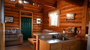 A log cabin interior with a kitchen area, a green door, a sofa, and a window with a curtain. - The Loon's Nest Cabin (Trapper Creek)