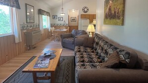 A spacious living area with a sofa, coffee table, and a rug. There is a kitchen in the background with wooden cabinets and a dining table. - Beaver's Hideaway Cabin (Trapper Creek)