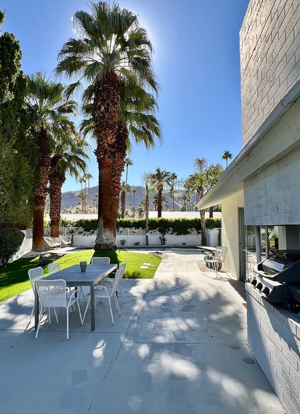 An outdoor patio area with a barbecue grill, white chairs, and palm trees.