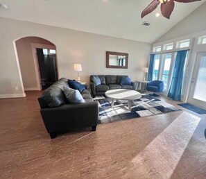 A spacious living room with a dark leather sofa, a round coffee table, and a large rug. There is a mirror on the wall and a door leading to another room.