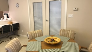 A dining area with a round table, checkered placemats, and a fruit bowl. There are four chairs with striped cushions. A clock and a thermostat are visible on the wall.