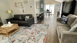 Un salón amplio con un sofá grande, una mesa de centro y una alfombra estampada. Al fondo, se aprecia un comedor con una mesa y sillas.