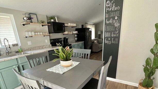 Una cocina moderna con zona de comedor, que presenta una mesa y sillas grises, una planta en maceta y una pizarra con mensajes de temporada.