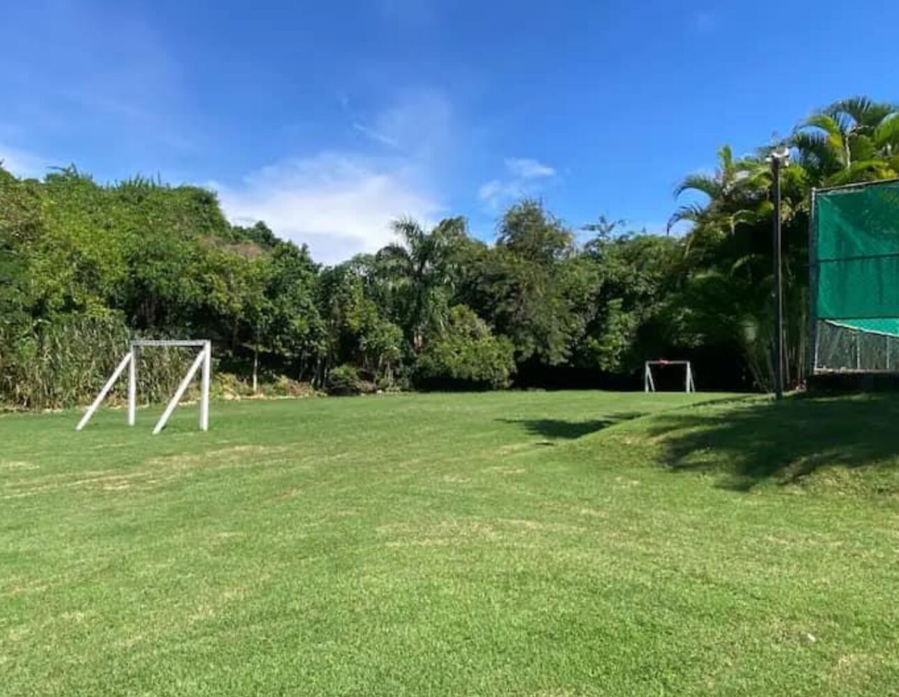 Un campo cubierto de césped con dos porterías de fútbol y una zona delimitada por una red verde, rodeado de árboles y un cielo despejado.