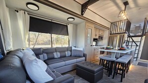 A modern RV interior with a sectional sofa, a dining area, and a kitchenette. - Fenced dog yard, creek fishing access, privacy and stunning sunset views! (Schoharie)