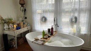 Una bañera independiente grande con artículos de aseo y una revista, una mesita con objetos decorativos, y un ventanal con cortinas translúcidas.