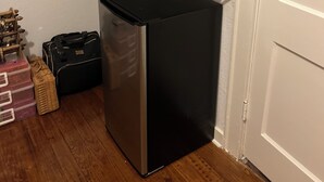 Una cocina compacta con un microondas en un estante, un refrigerador y recipientes de almacenamiento en el suelo.
