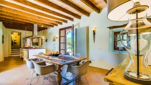 A spacious dining area with a wooden table and chairs, a kitchen in the background, and a large window with a view of greenery. - Cozy home in Llubi with private swimming pool, can be inside or outside (Llubi)