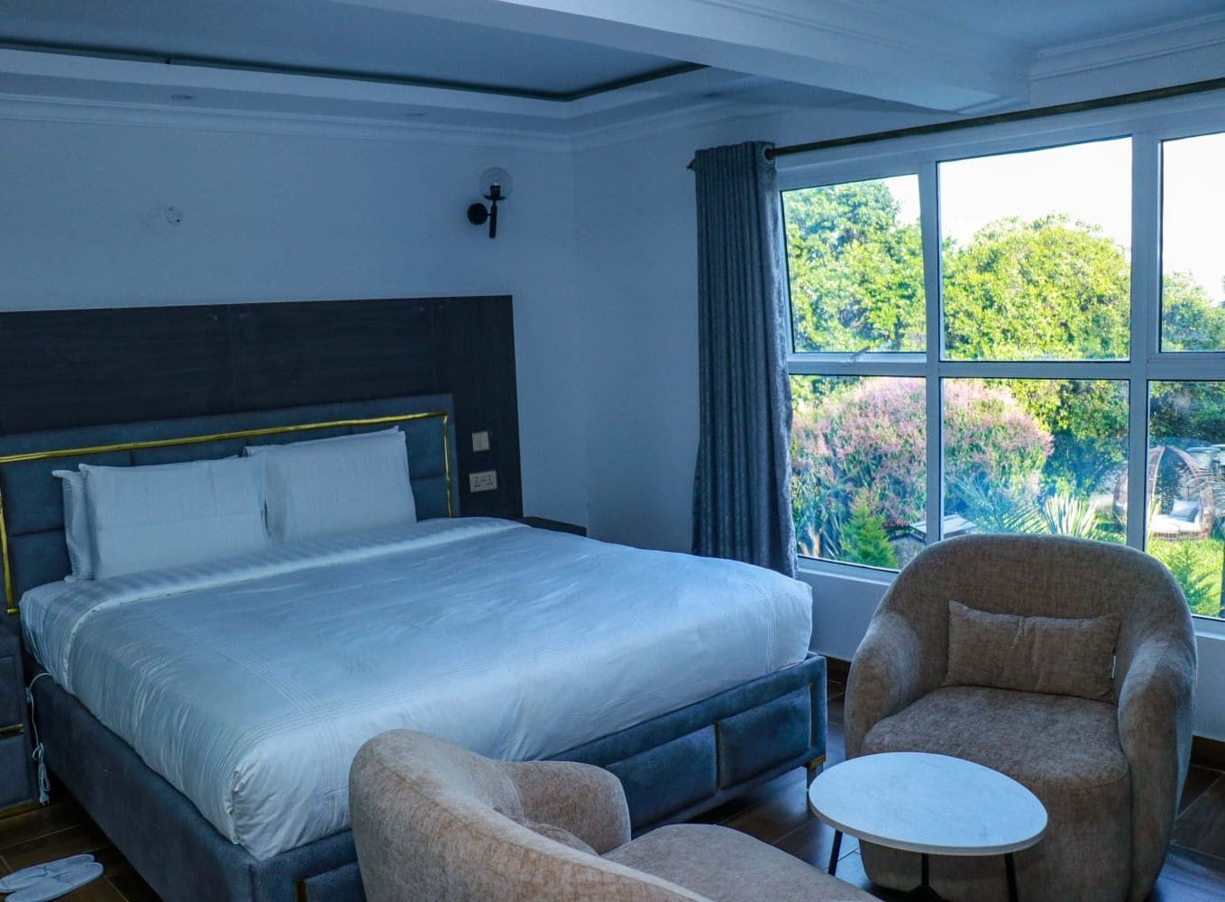 A hotel room with a large bed, a chair, a small table, and a window with a view of greenery.