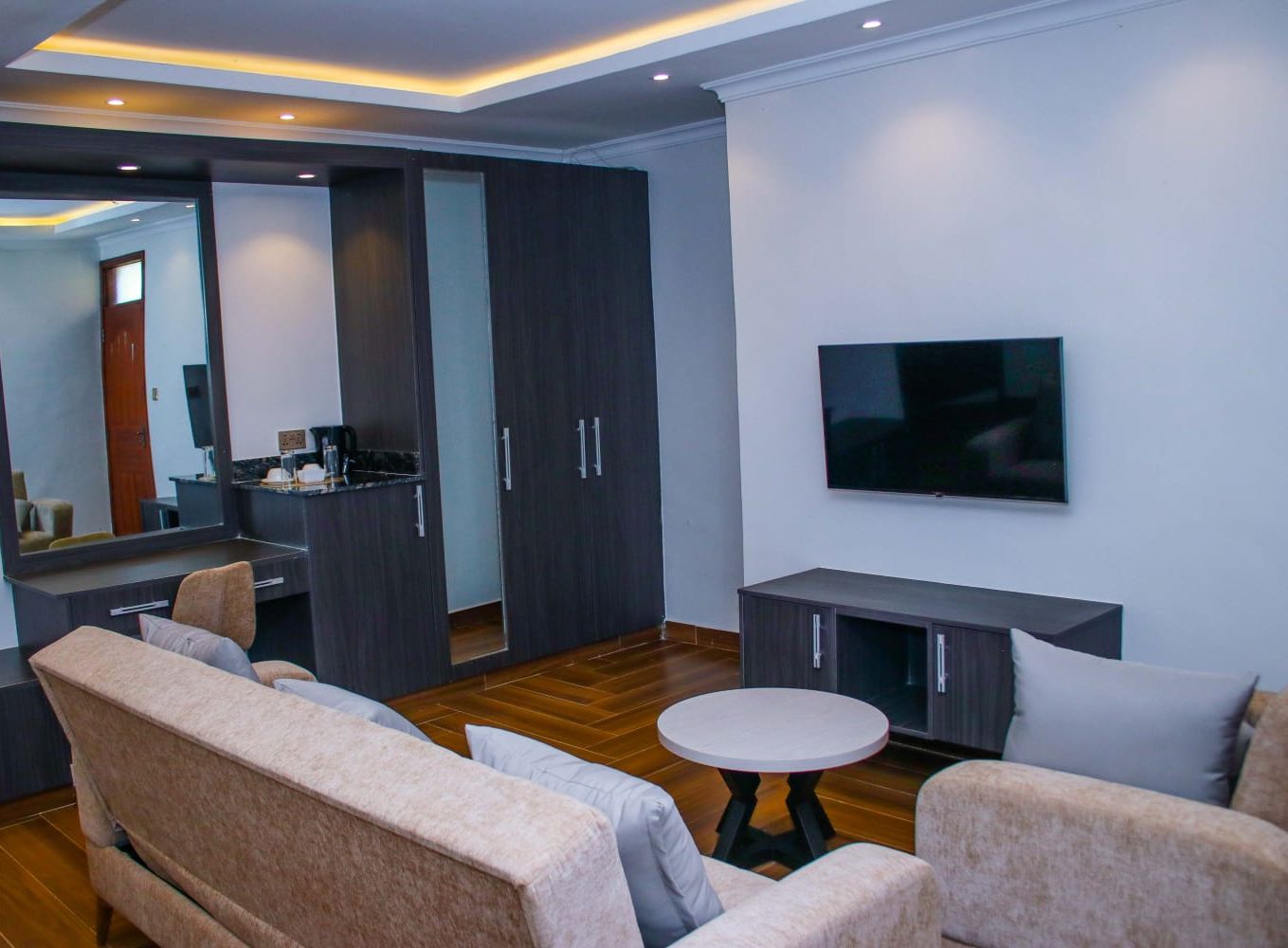 Ein modernes Wohnzimmer mit einer Couch, einem runden Couchtisch, einem an der Wand montierten Fernseher und einer Küchenzeile mit Schränken und einer Arbeitsplatte.