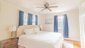 Une chambre à coucher avec un lit, une table de chevet, un ventilateur de plafond et deux fenêtres avec des rideaux bleus.