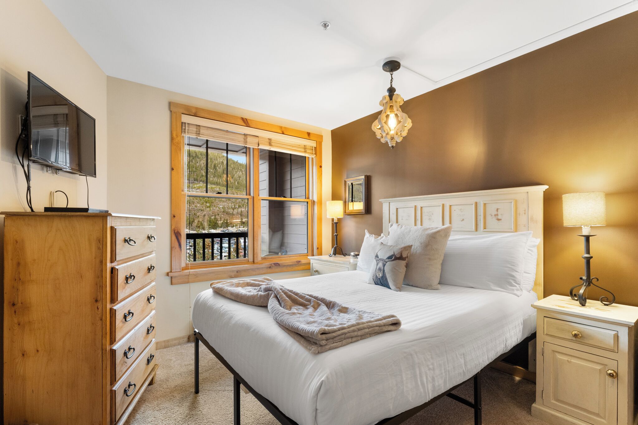 A bedroom with a bed, a dresser, a nightstand, a lamp, a TV, and a window with blinds.