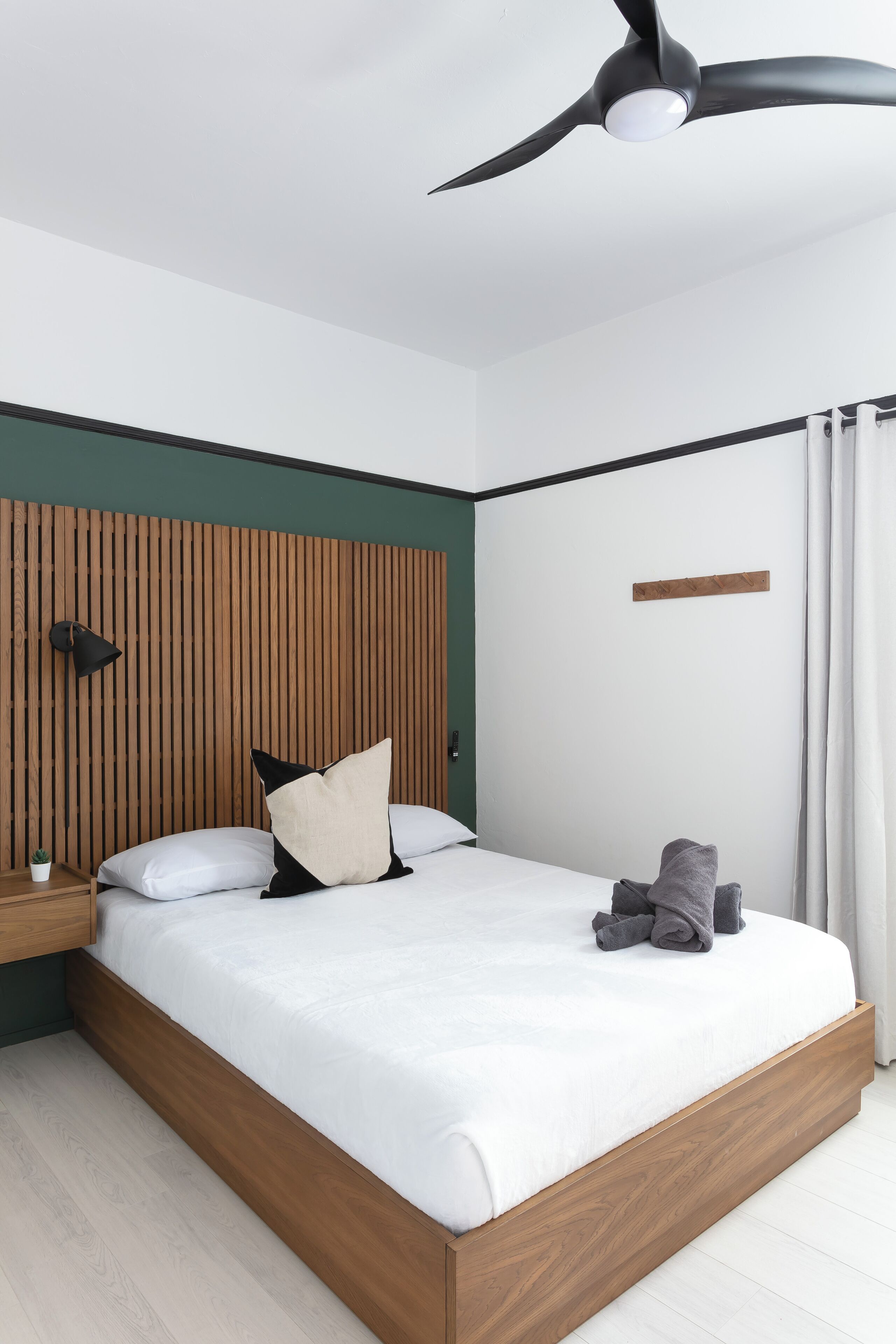 A modern bedroom with a wooden headboard, a ceiling fan, a bedside table with a lamp, and a neatly made bed with pillows and a blanket.