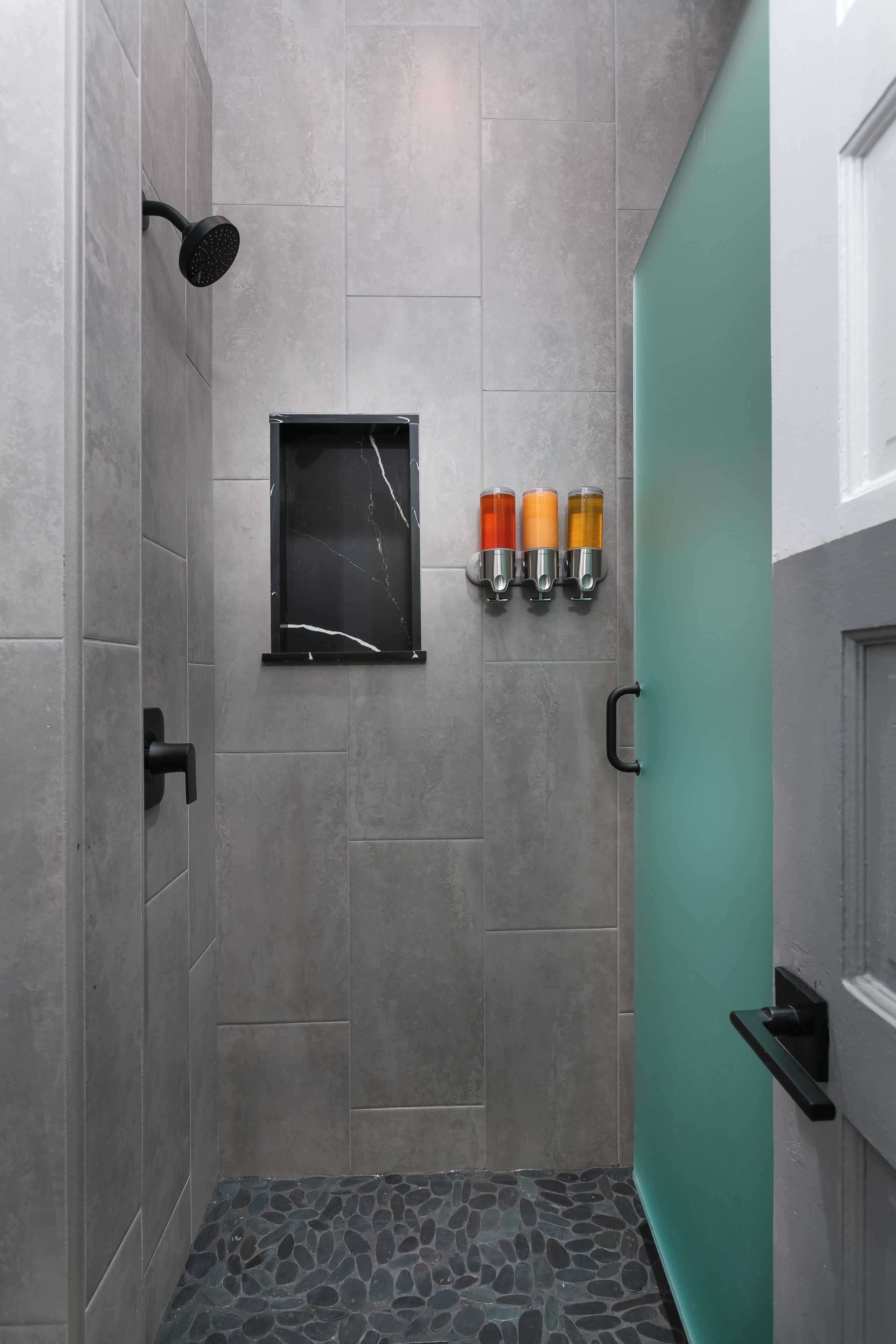 A modern shower area with grey tiles, a black showerhead, a glass door, and three orange bottles on the wall.