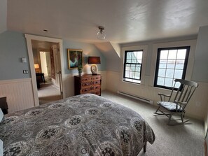Un dormitorio con una cama grande, una mecedora, un aparador, un cuadro y una ventana con vistas.