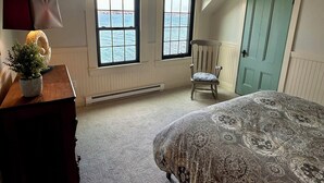 Ein Schlafzimmer mit Bett, Stuhl, Kommode, Lampe und Meerblick durch zwei Fenster.