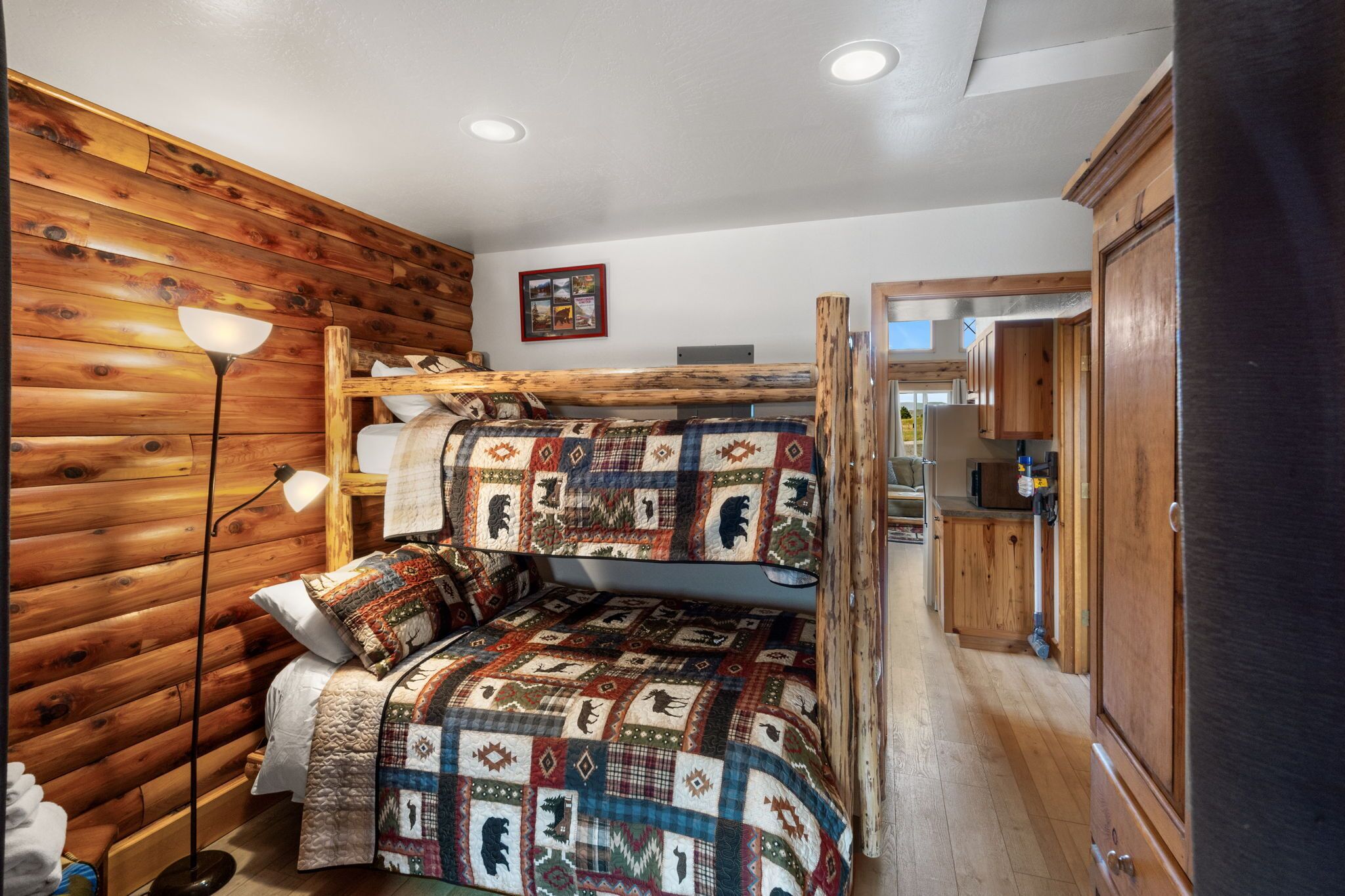 A cozy cabin-style room with a bunk bed, wooden walls, a quilted bedspread, a floor lamp, and a wooden cabinet.