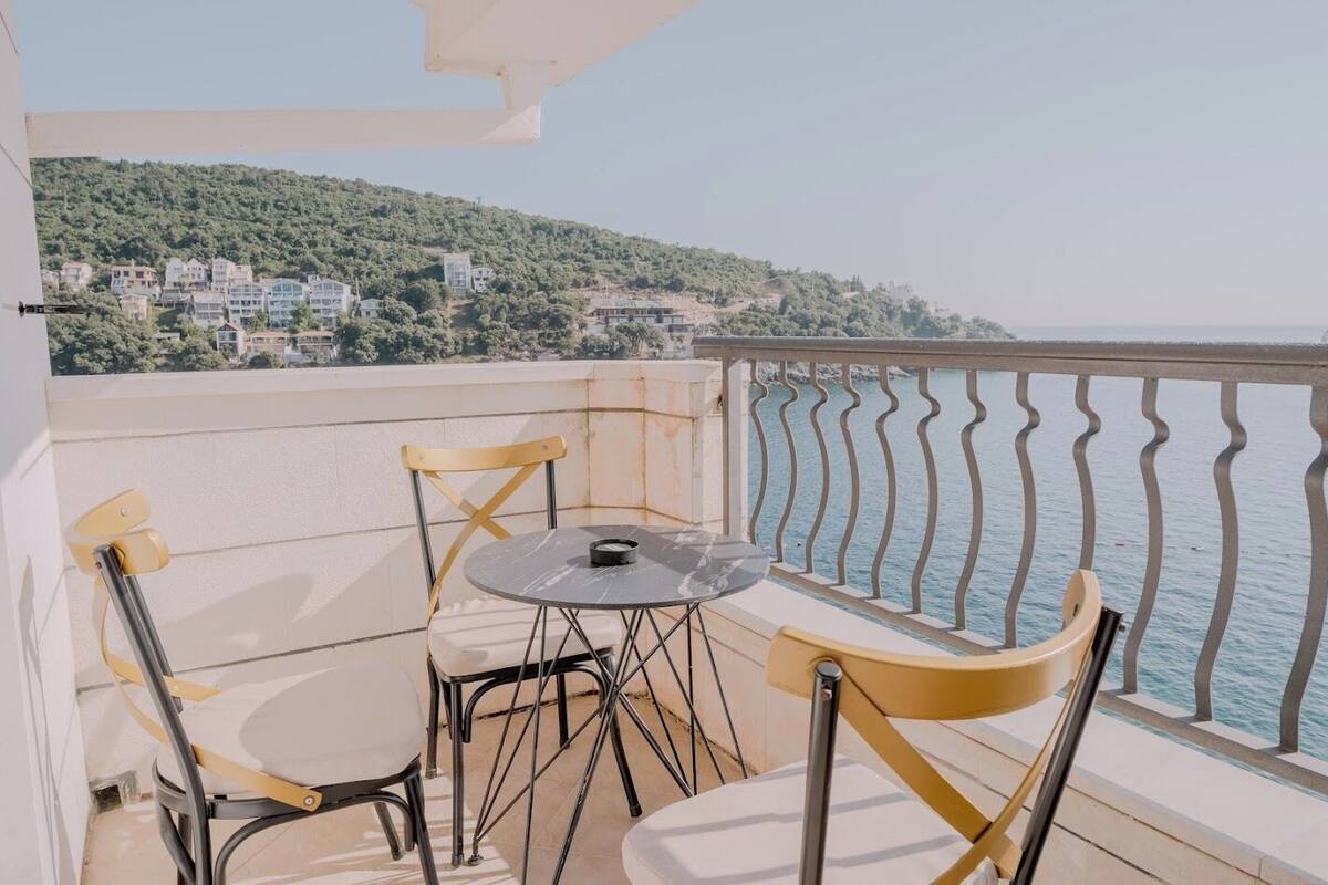 A balcony with a table and chairs, overlooking a body of water and a hillside with buildings.