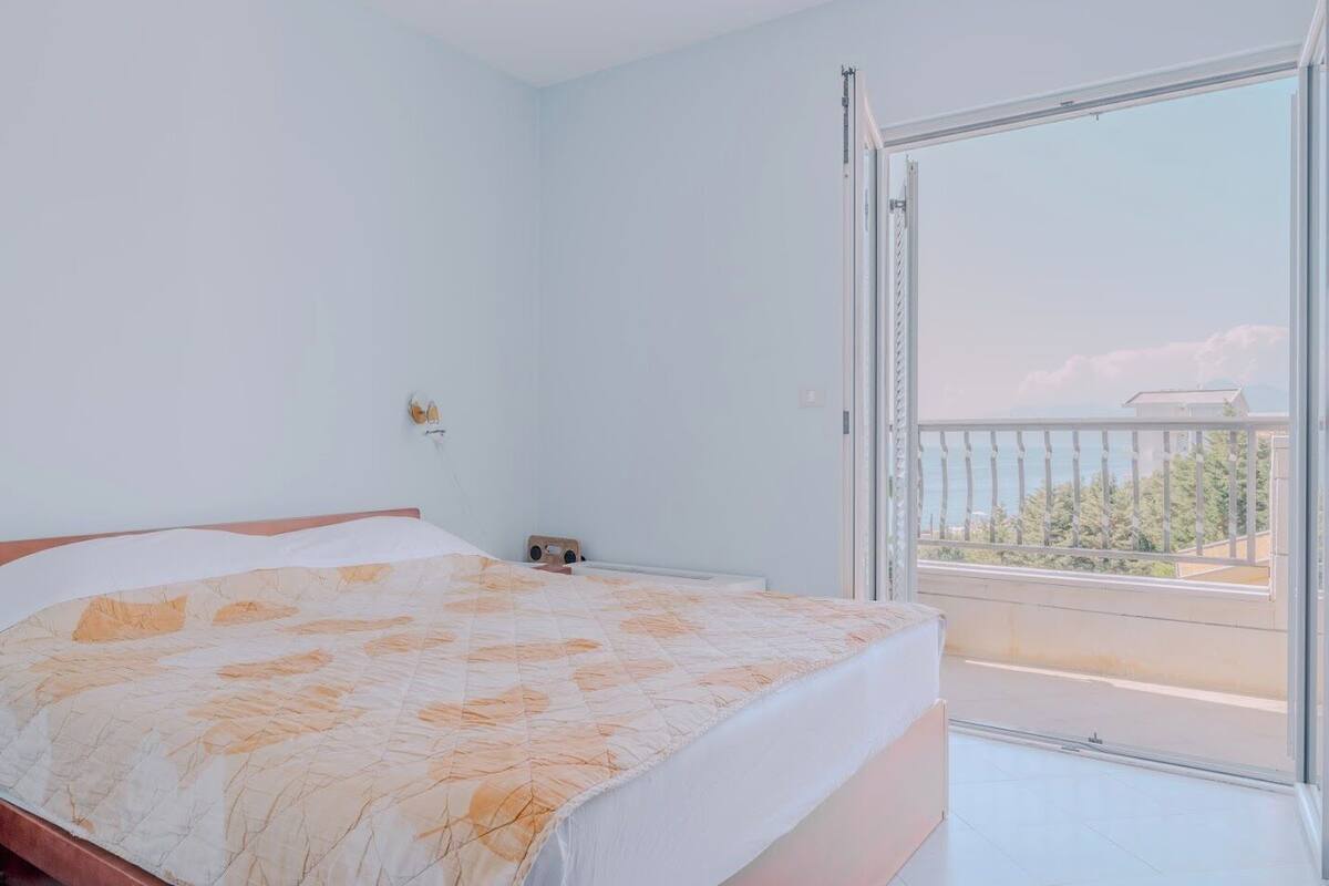 A bedroom with a bed, a small bedside table, a wall socket, and a balcony with a view of the sea and trees.