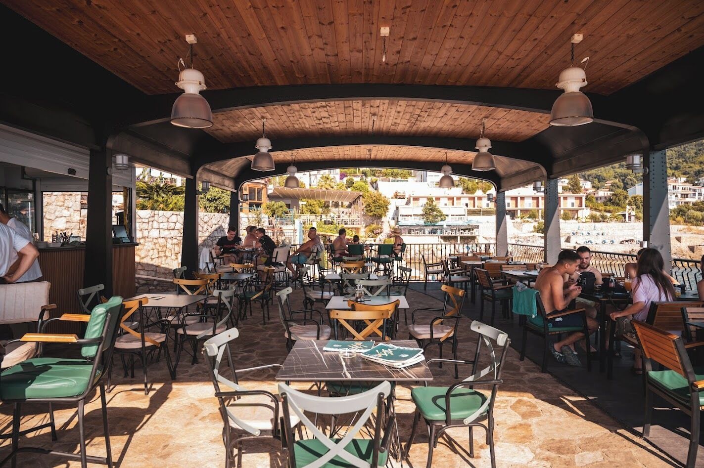 An outdoor café with wooden tables and chairs, a stone floor, and a view of buildings in the background.