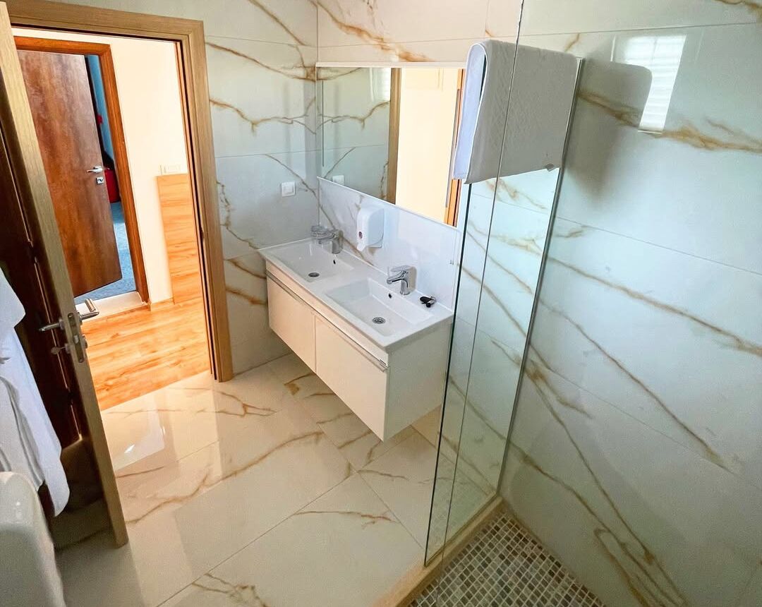 A bathroom with a marble floor, a double sink vanity, a glass shower enclosure, and a wooden door.