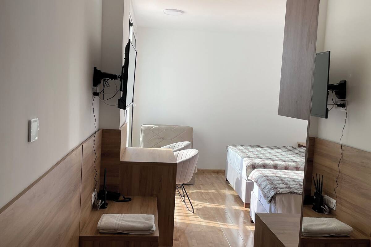 A hotel room with a wooden floor, a bed with a plaid duvet, a chair, a desk, and a mirror.