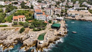 Luftaufnahme einer Küstenstadt mit felsiger Küstenlinie, einem Yachthafen und einem Strandabschnitt mit Liegestühlen und Sonnenschirmen.