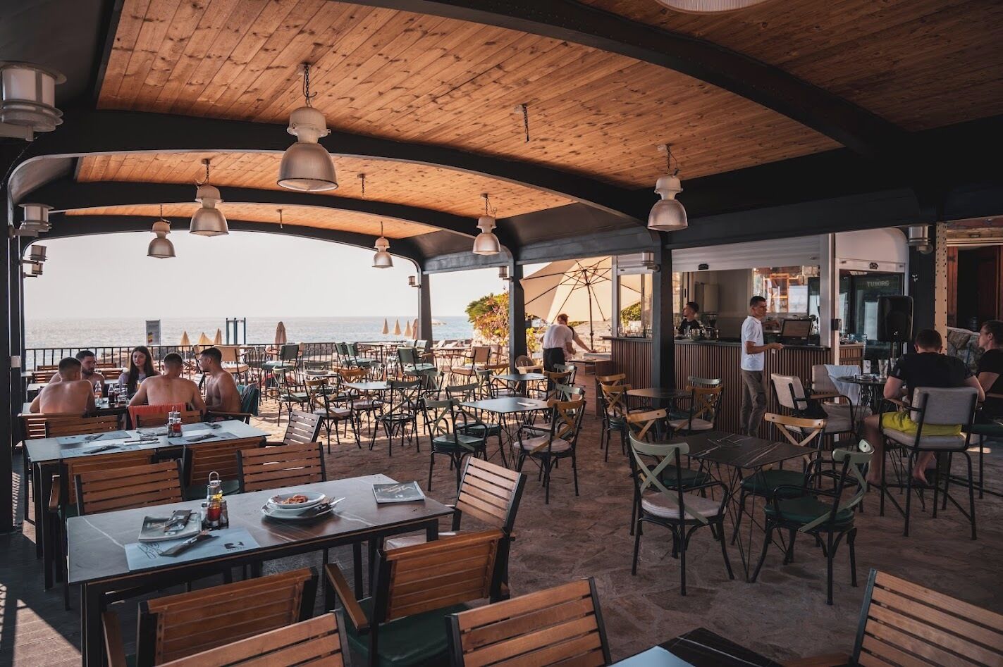 An outdoor dining area with wooden tables and chairs, a bar area, and a view of the sea.